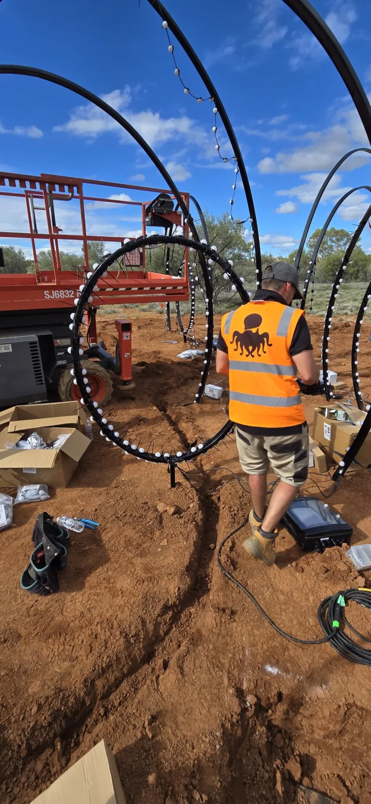 An installer in a hi-vis vest fitting pixel dots to a hoop, a scissor lift parked behind