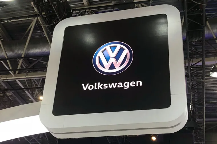 白色圆角框上的 Volkswagen 发光标识，从下方仰拍，背景是展馆的桁架