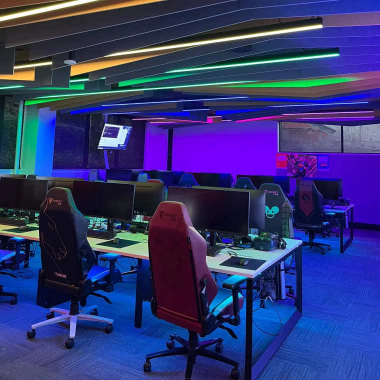 Rows of desks and gaming chairs under angled ceiling battens washing green, blue and violet across the room