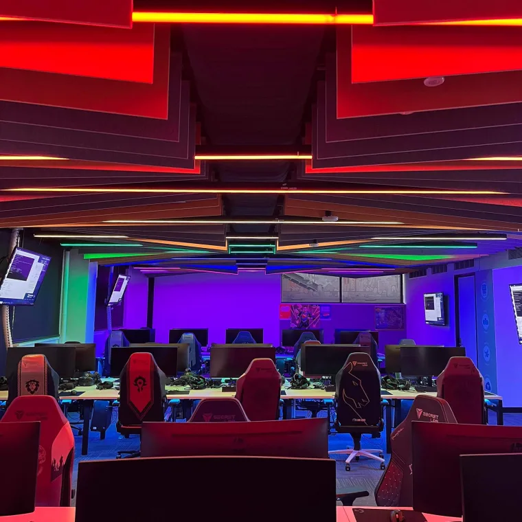 Red and gold light across the ceiling with rainbow battens above empty red gaming chairs, looking down the length of the room