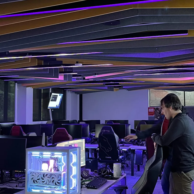 A student leaning over a desk to adjust a PC, ceiling battens in pink and violet above the workstations