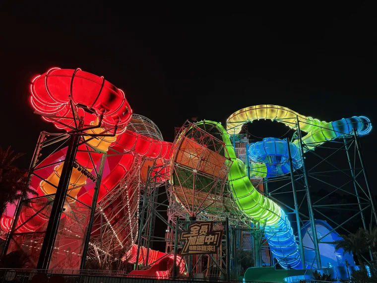 The tower complex lit red on one side while a green and blue tube snakes away past illuminated Chinese signage