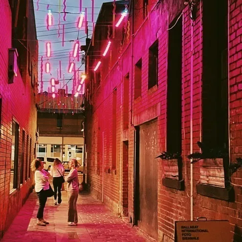 三位好友走在 Hop Lane 的灯光顶棚下,洋红色光洒在砖墙上,墙面挂着 Ballarat International Foto Biennale 的标识