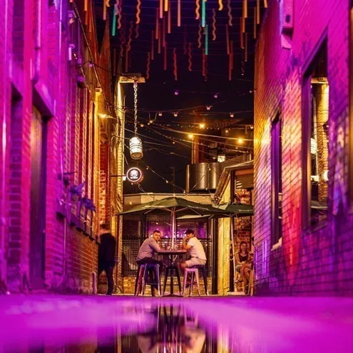 深夜的 Hop Lane,两人站在高脚桌旁,头顶的粉色与橙色灯光映在湿润的巷道地面上