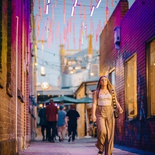 暮色中的 Hop Lane,一位身着米色服装的女子朝镜头走来,身后是伞下的人群,头顶悬挂着彩色灯管