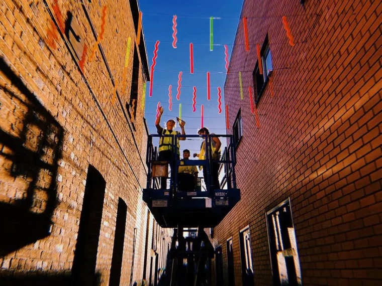 两名身穿反光衣的安装人员站在升起的剪叉式升降平台上,在 Hop Lane 的砖墙之间安装彩色亚克力条,背景是蓝天