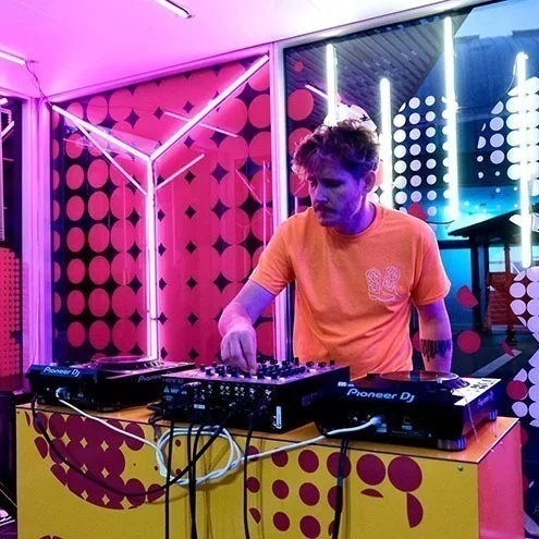 一位身穿橙色 T 恤的 DJ 在 Boom Box 内操作打碟机,身后是粉黑波点面板