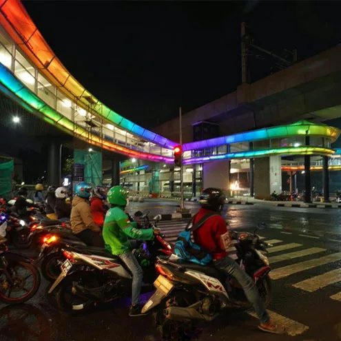 摩托车骑手在弧形步道下的路口等待，连廊底部被橙色、绿色与蓝色灯光洗亮
