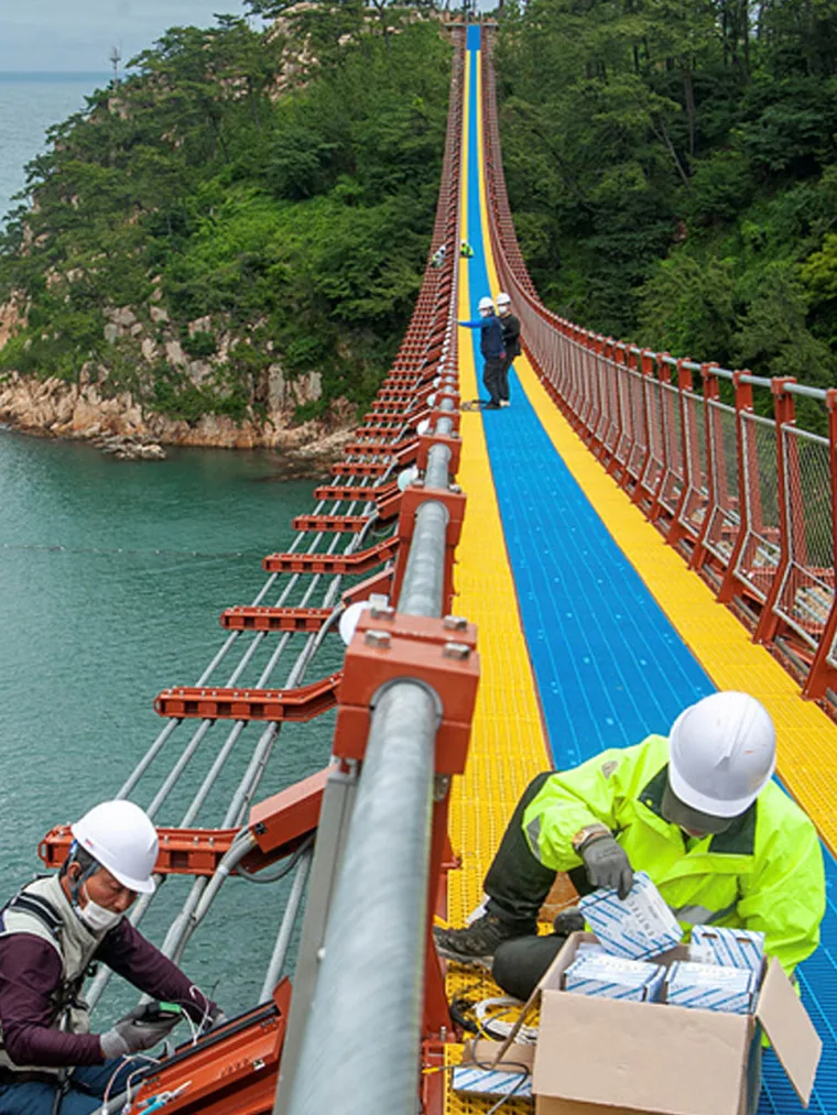 两名戴白色安全帽、穿反光背心的工人跪在桥面上，从纸箱中取出灯具进行安装，身后是绿色海岬