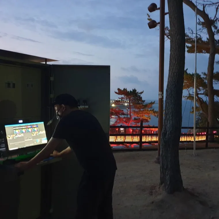 黄昏时分，一名技术人员在控制柜前操作显示器，身后松林中的桥体泛着红色与琥珀色的光