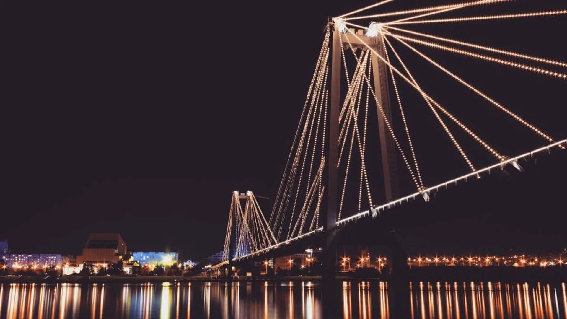 夜间桥体呈暖琥珀色与白色，桥跨下方是城市灯光与水面上长长的倒影