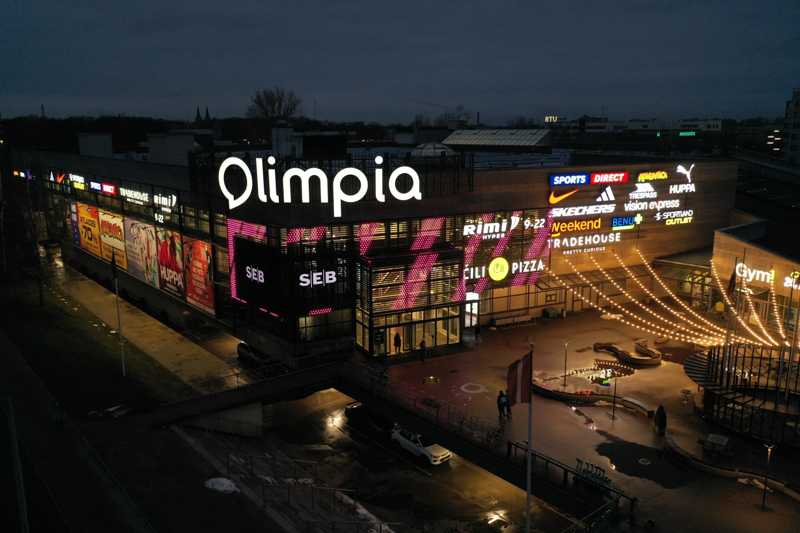 Pink blocks of colour running across the facade, shoppers crossing the plaza under the festoon lights