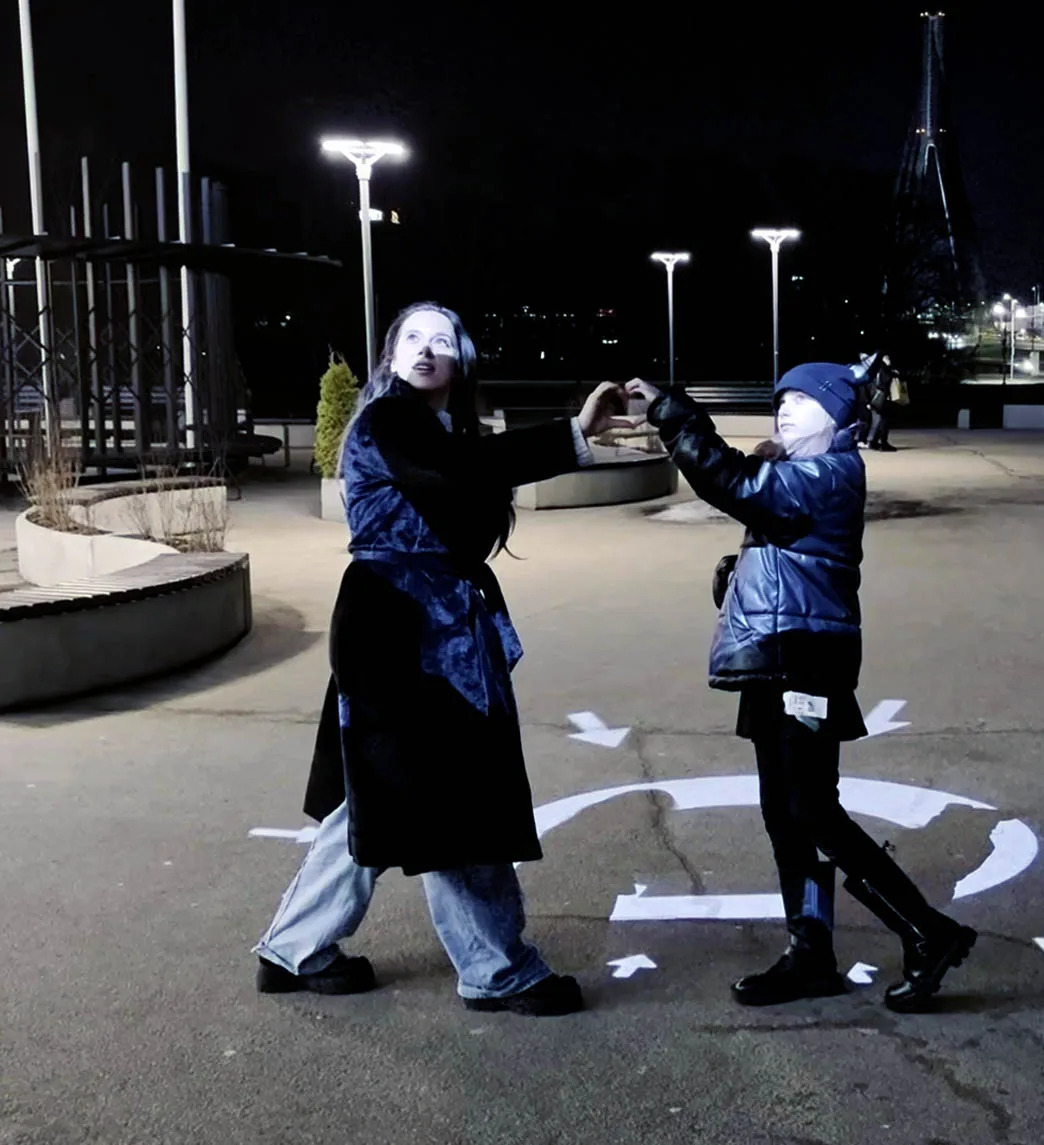 Two people making a heart with their hands on a lit circular marker in the plaza at night, arrows pointing in around them