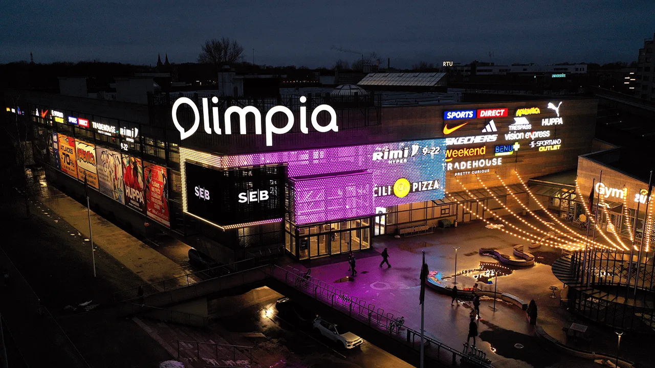 Olimpia shopping centre at dusk, its LED facade glowing magenta above a plaza strung with warm festoon lights