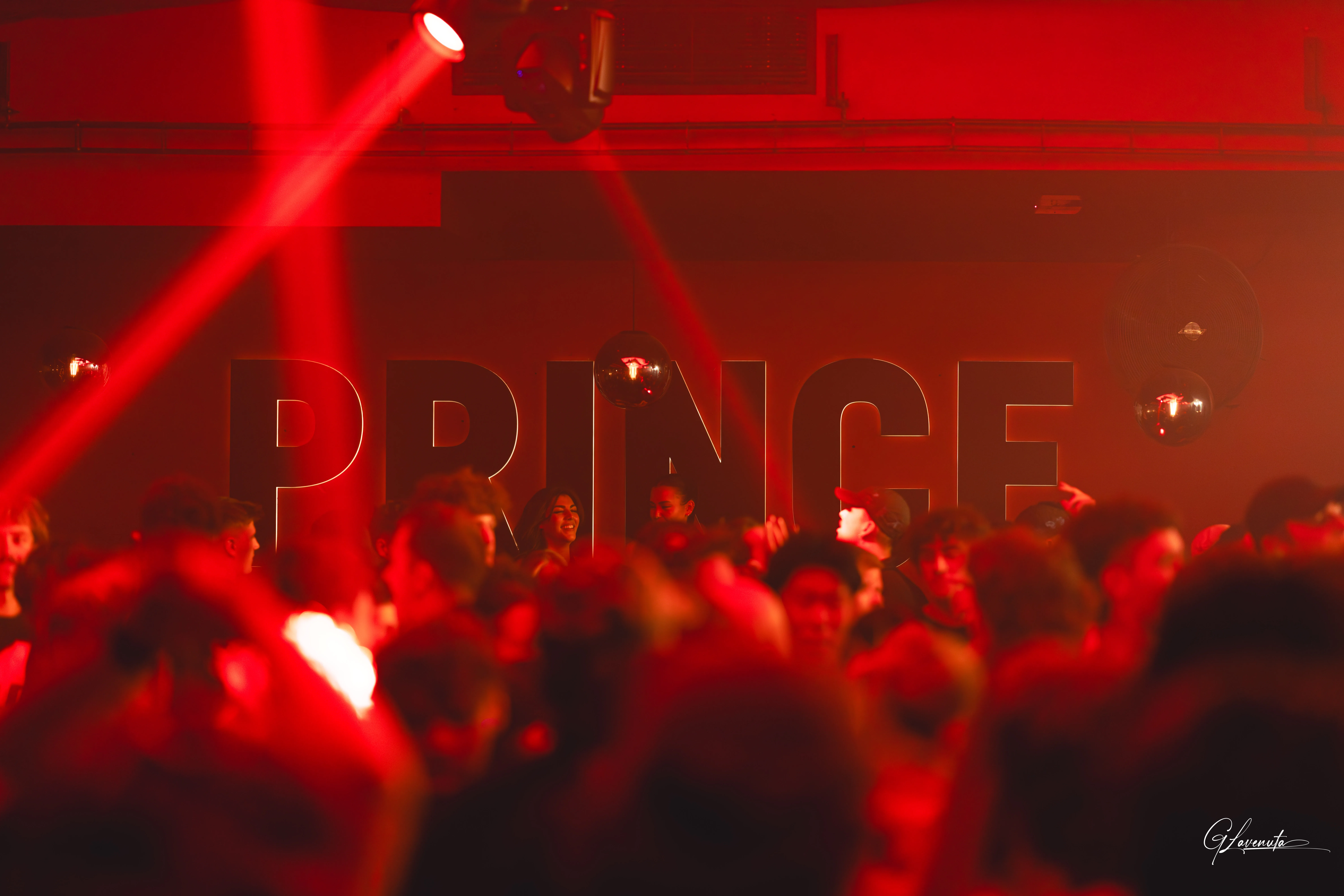 红色光束穿透人群上方的烟雾,后墙上的 PRINCE 字样被染成红色