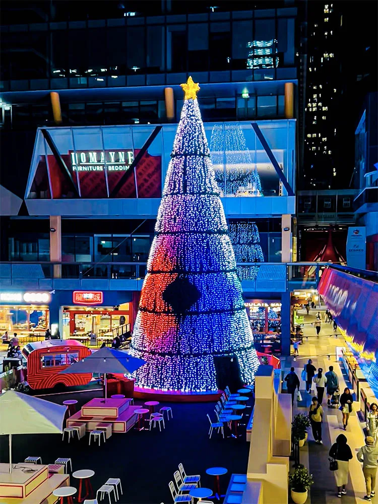 从上层平台俯瞰圣诞树,锥体依次呈现绿、蓝、白色,下方人群在美食摊位间穿行