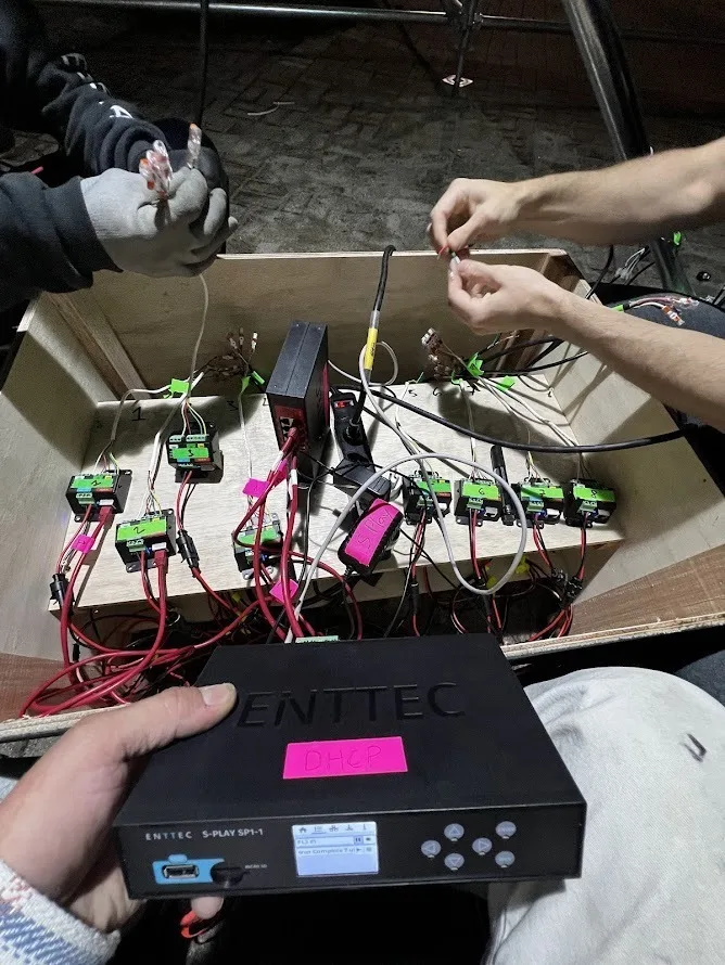 现场，一双手正在为一箱绿色 PLink Injector 接线，前方举着一台 ENTTEC S-PLAY SP-1 控制器