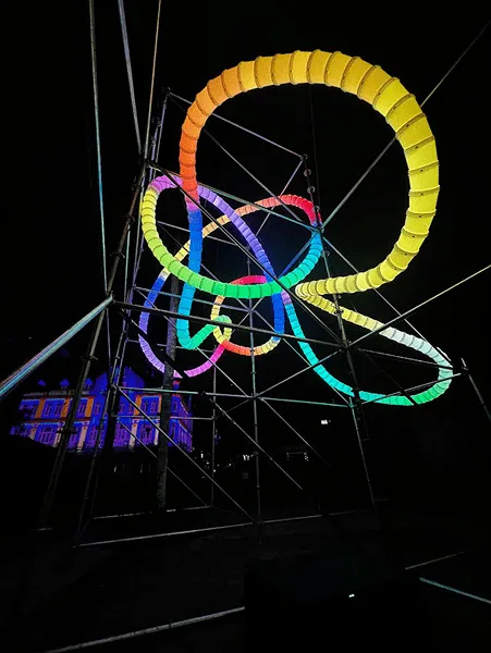 夜间黑色脚手架框架内的环形装置亮起黄、绿、蓝、粉色，拉索在黑暗背景中交错