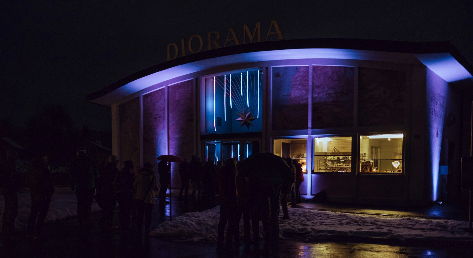 夜色中 Museum DIORAMA 的弧形玻璃入口,蓝色与紫色光线透过窗户洒出,参观者聚集在门外