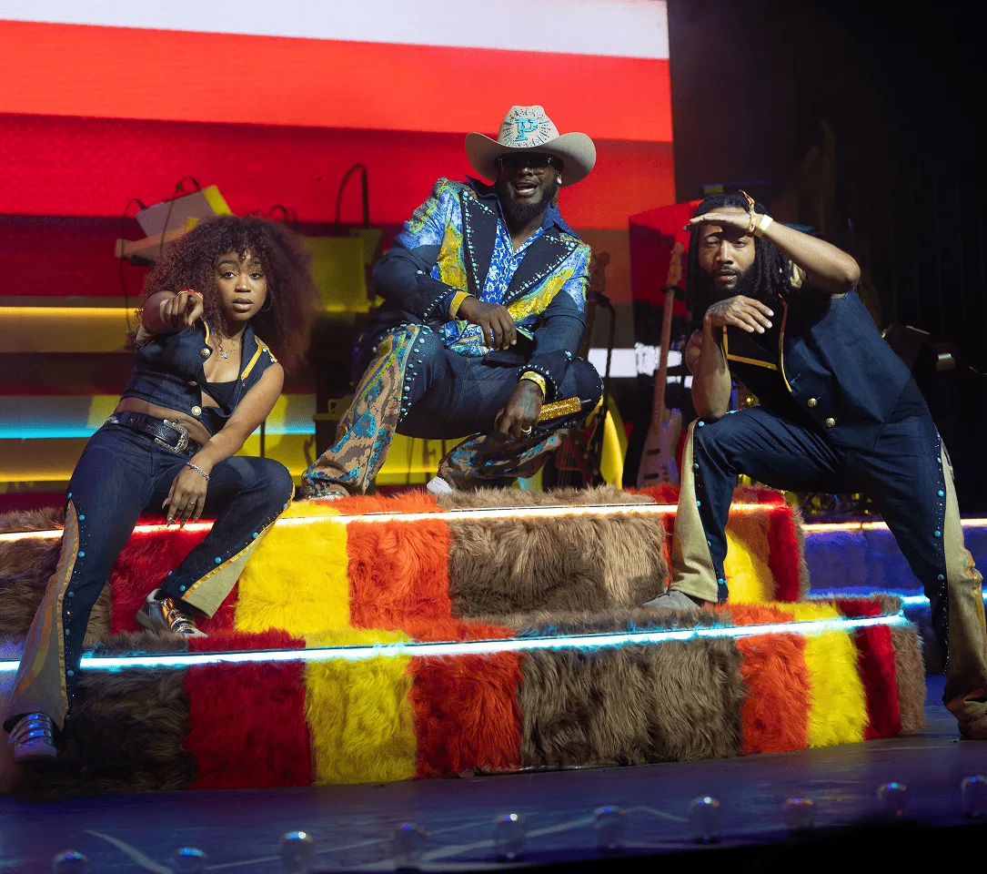 Three performers posed across the shag-carpet steps, the central figure in a white hat and sequinned blue suit