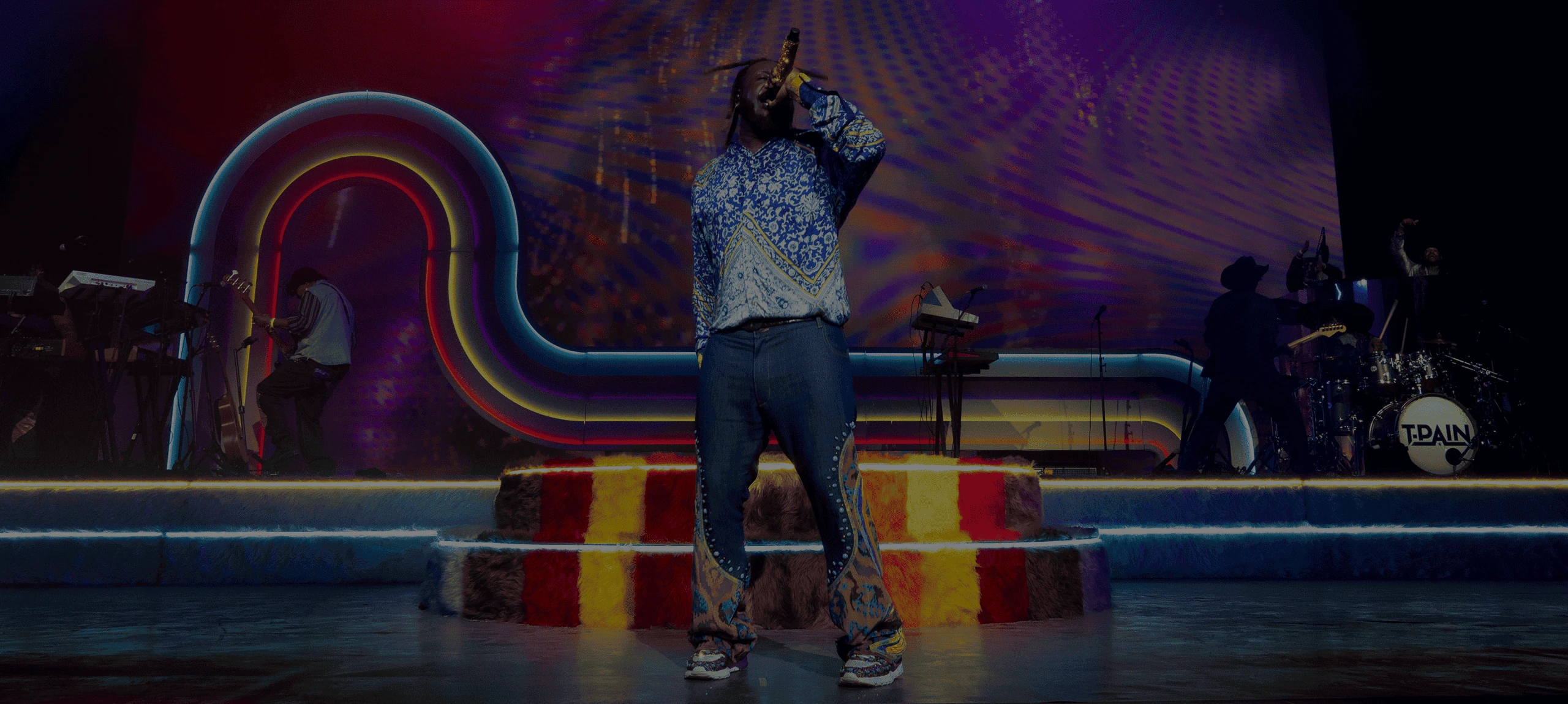T-Pain seen from behind with his arms raised, the swooping neon arch and shag-carpet platform lit around him on the Palomino Stage