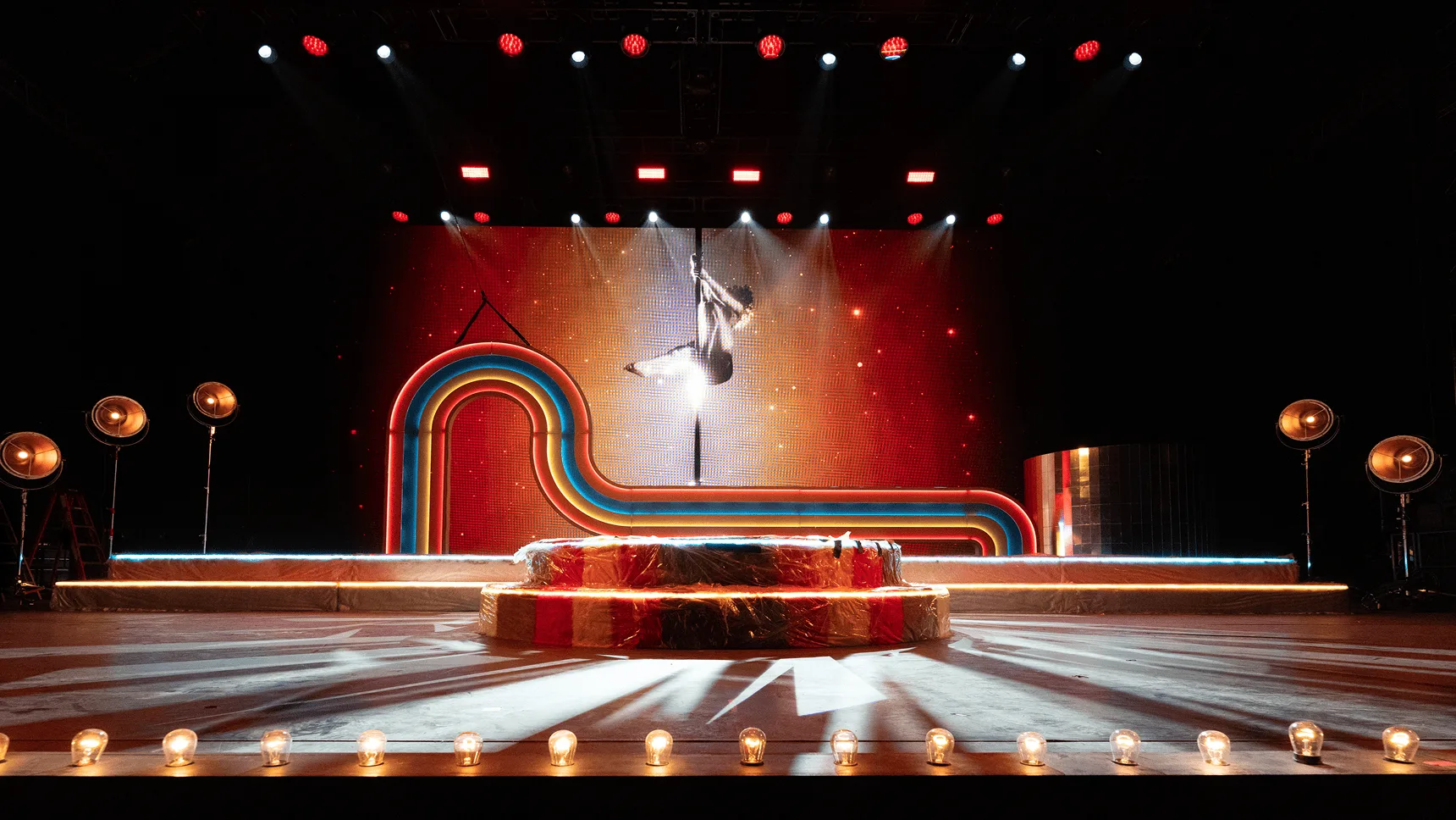 The empty set before doors, an aerialist descending over the thrust with warm footlights running along the stage edge