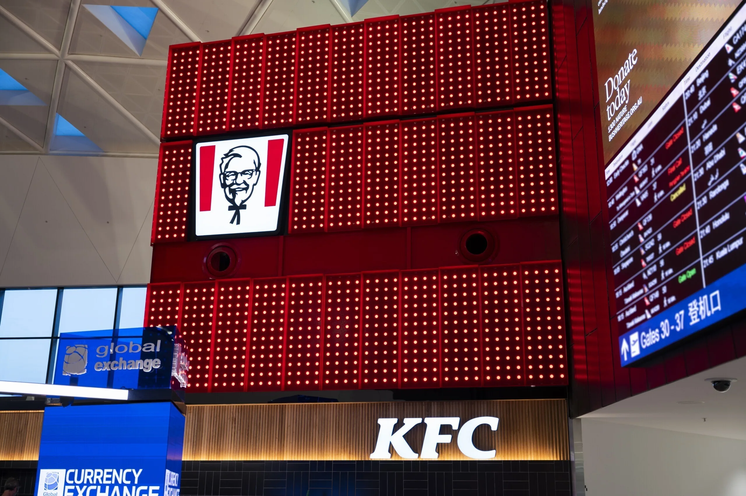 灯盒转角上是白色 KFC 字标，下方是航班信息屏与货币兑换柜台