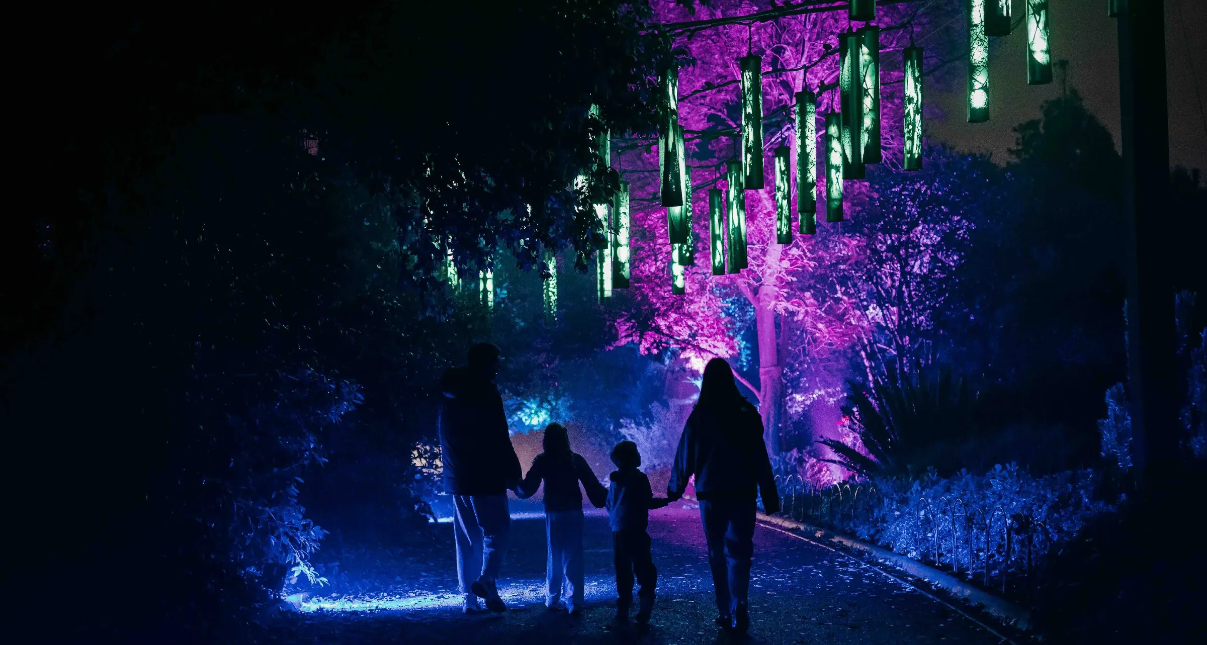 A family silhouetted on the garden path at night, walking beneath the lit lanterns of WILDERNESS WITHIN